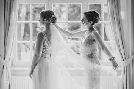 Braut mit Trauzeugin am Fenster bei der Vorbereitung zur Hochzeit